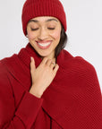 A smiling woman wearing a matching ribbed knit beanie and a thick ribbed knit wrap or scarf in a deep red color, posed against a light gray studio background.