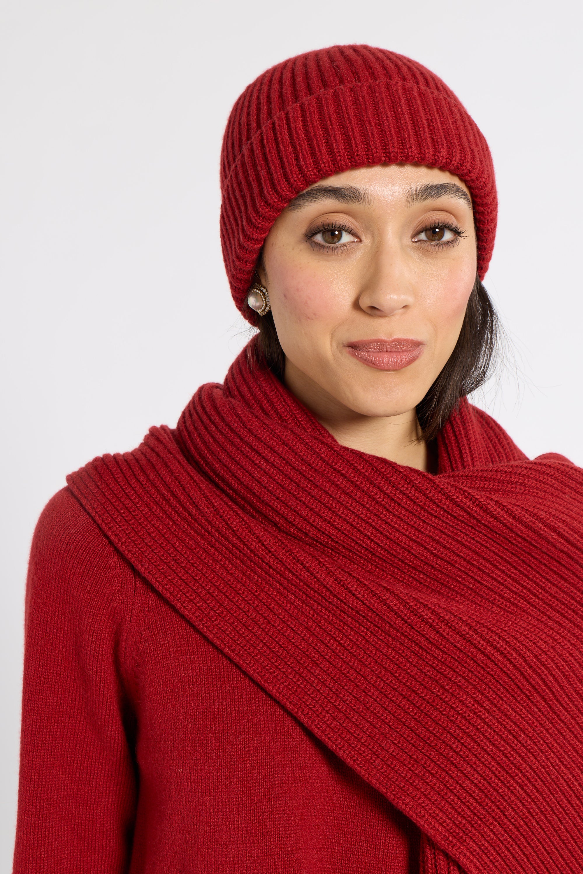 A woman wearing a matching red ribbed knit beanie and a wide red wrap scarf, photographed against a plain light gray studio background.