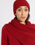 A woman wearing a matching red ribbed knit beanie and a wide red wrap scarf, photographed against a plain light gray studio background.