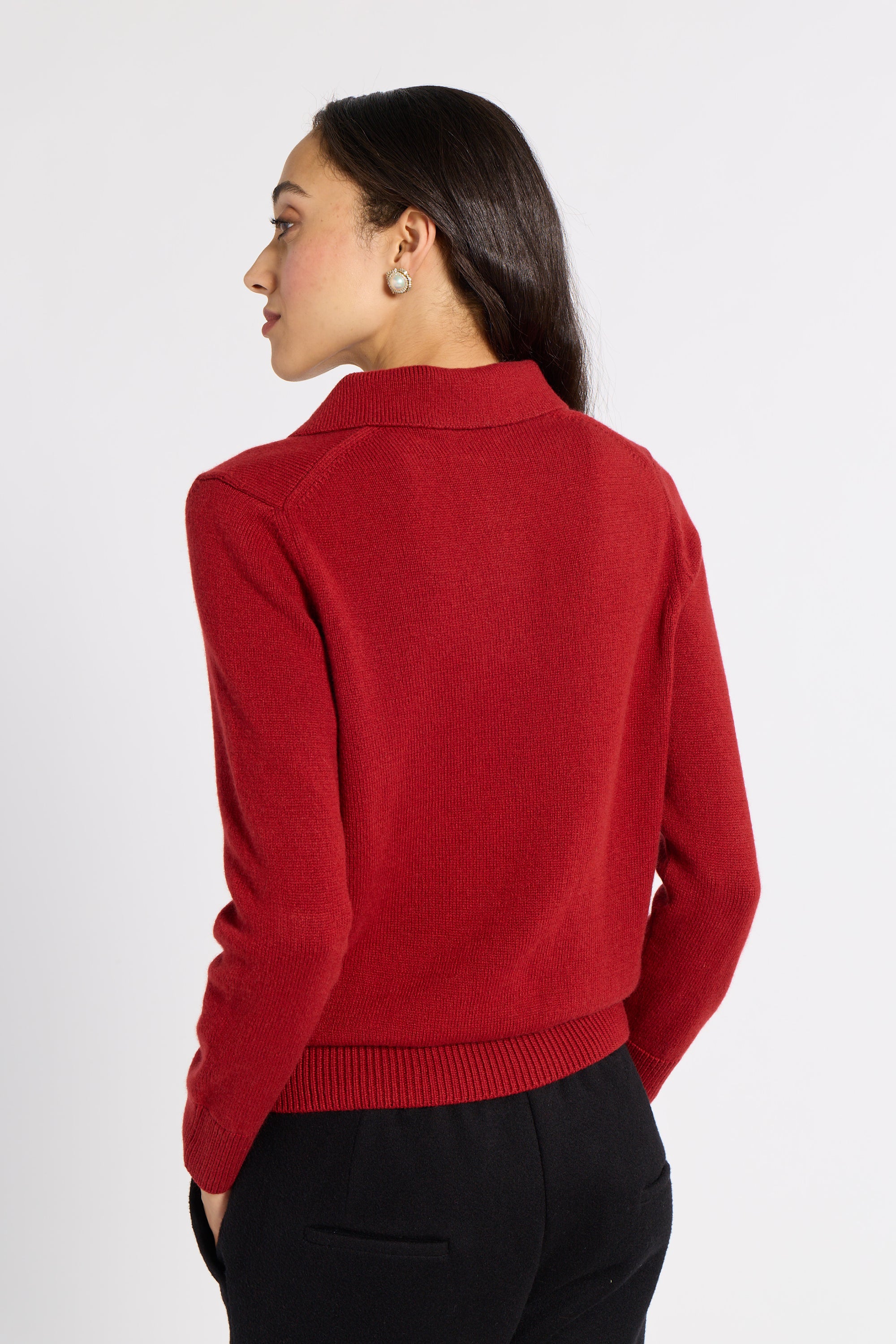A woman shown from the back wearing a red knit sweater with a mock neck and black trousers, photographed against a plain light gray studio background.