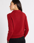 A woman shown from the back wearing a red knit sweater with a mock neck and black trousers, photographed against a plain light gray studio background.