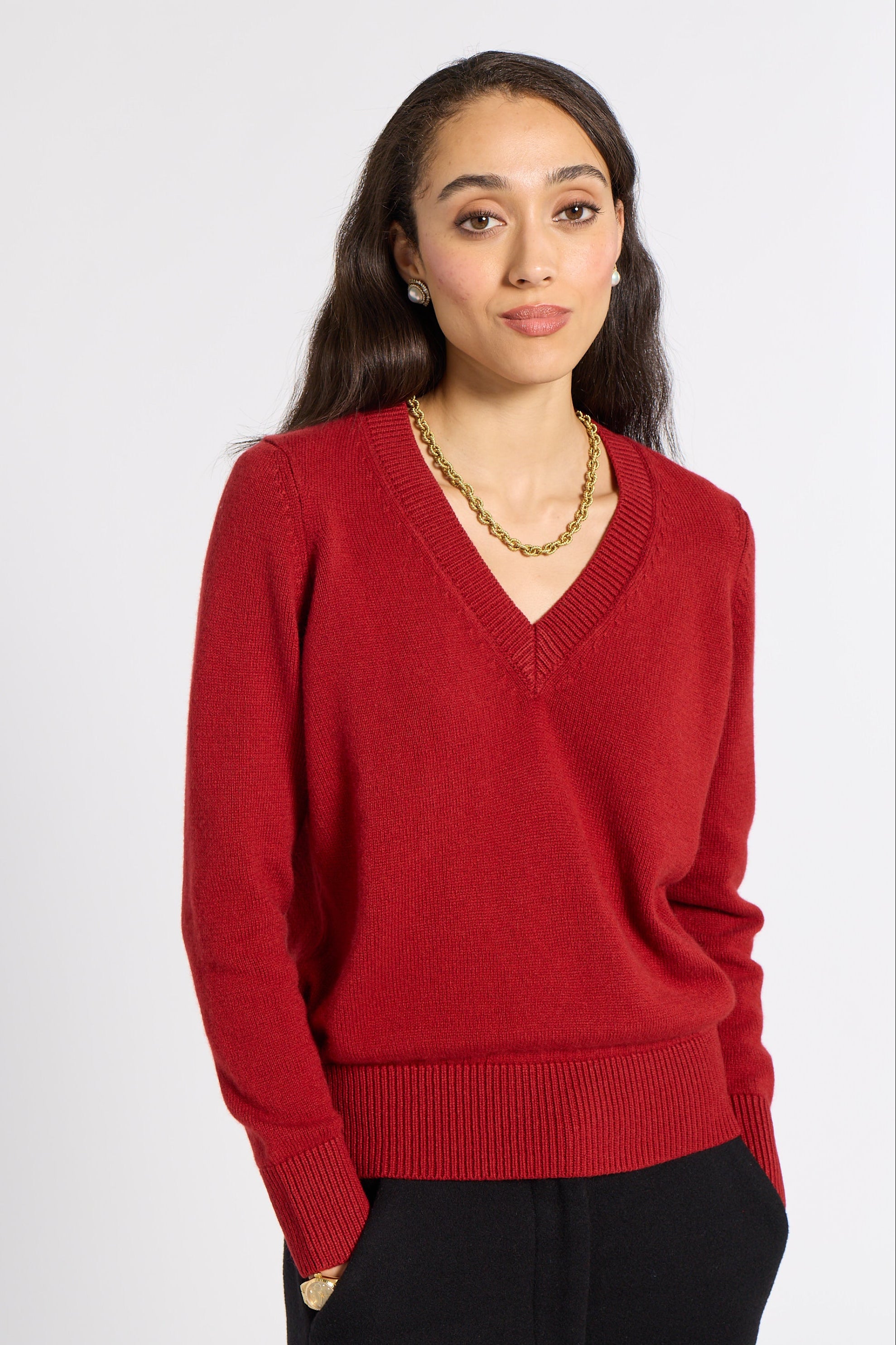 A woman modeling a red V-neck knit sweater with black trousers, standing against a plain light gray studio background.