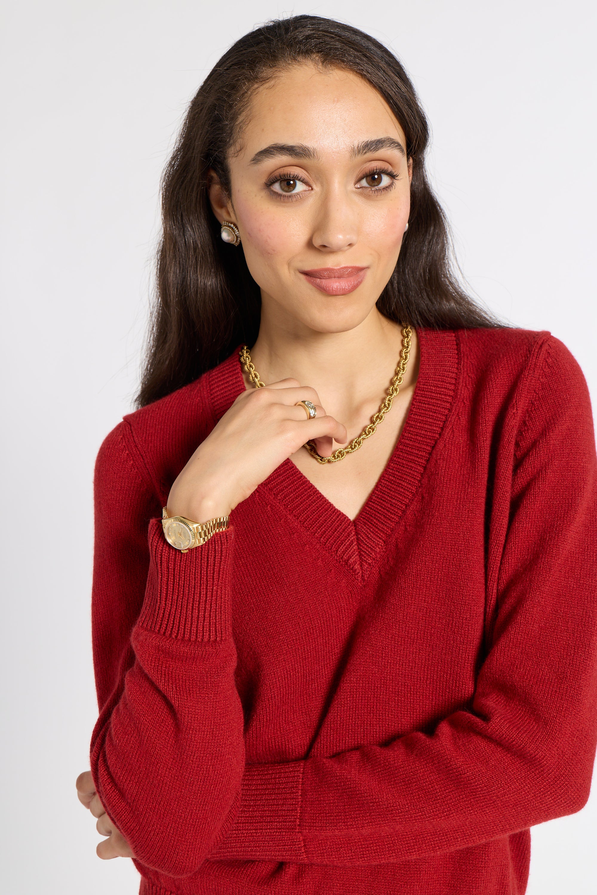 A woman wearing a red V-neck knit sweater, styled with a gold chain necklace, gold watch, and gold ring, against a light gray studio background.