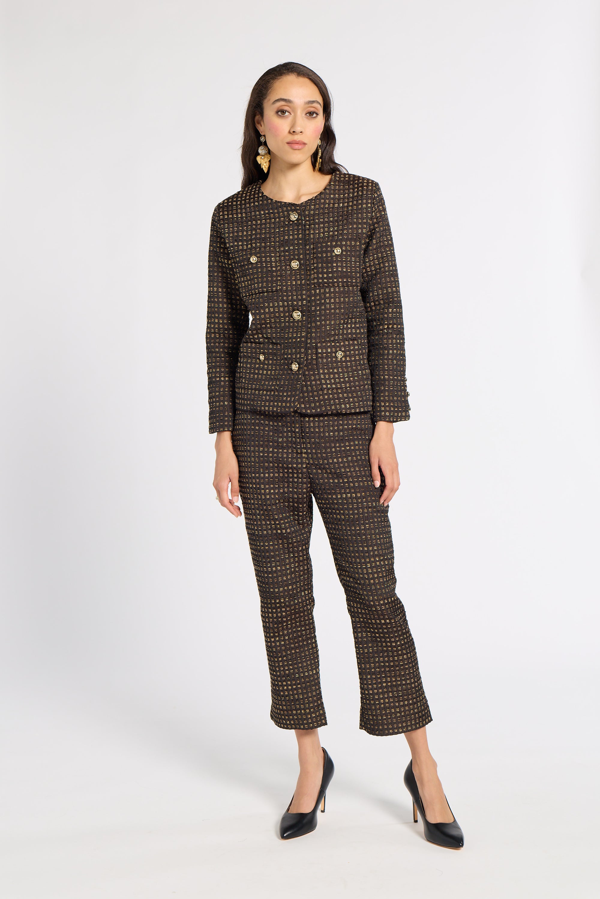 A woman modeling a matching brown tweed jacket and cropped trousers set with gold-tone buttons, paired with black pointed-toe heels, against a plain light gray studio background.