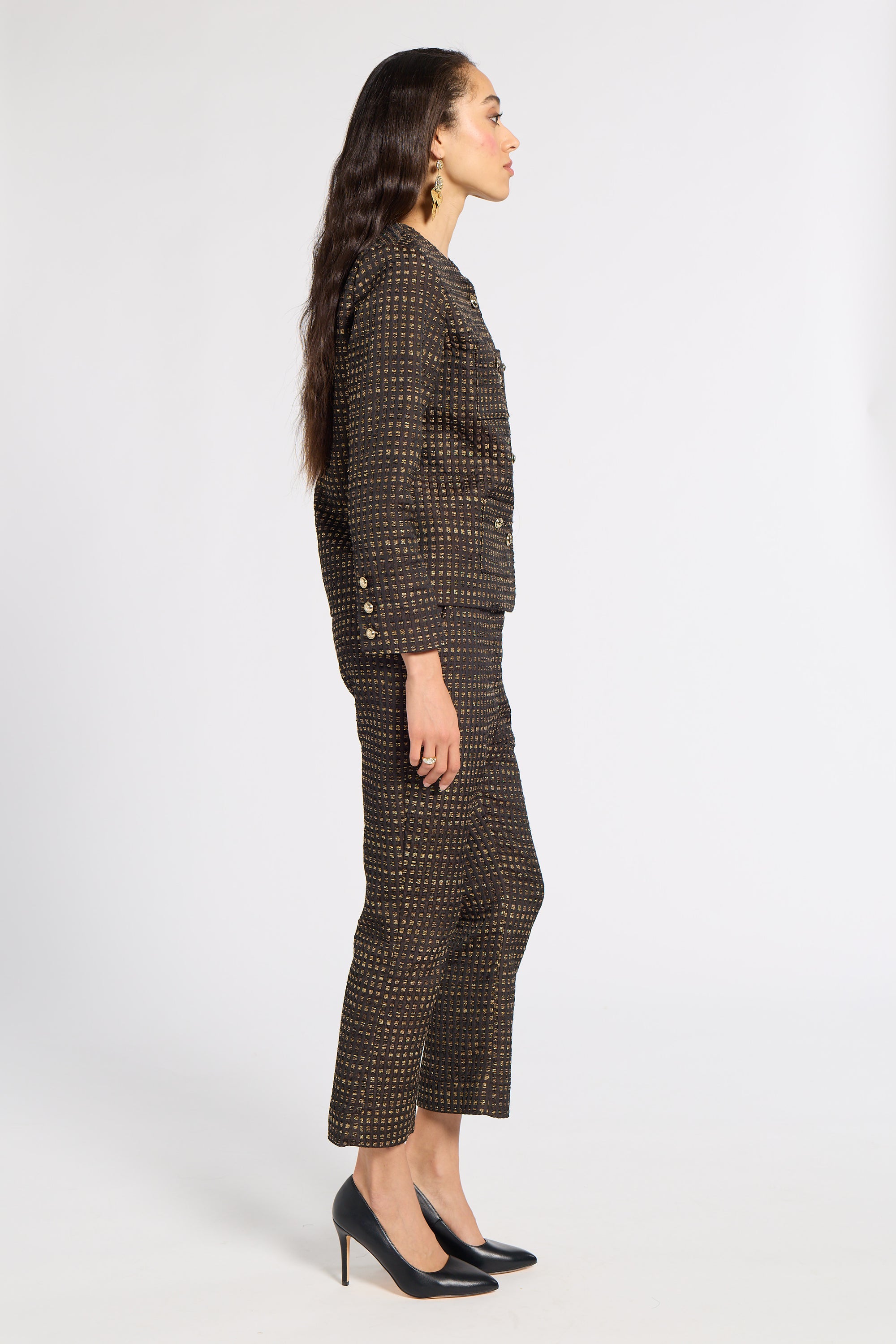 A woman stands in profile wearing a dark tweed suit with a fine gold geometric pattern, paired with black pointed-toe heels, against a plain light gray studio background.
