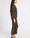 A woman stands in profile wearing a dark tweed suit with a fine gold geometric pattern, paired with black pointed-toe heels, against a plain light gray studio background.