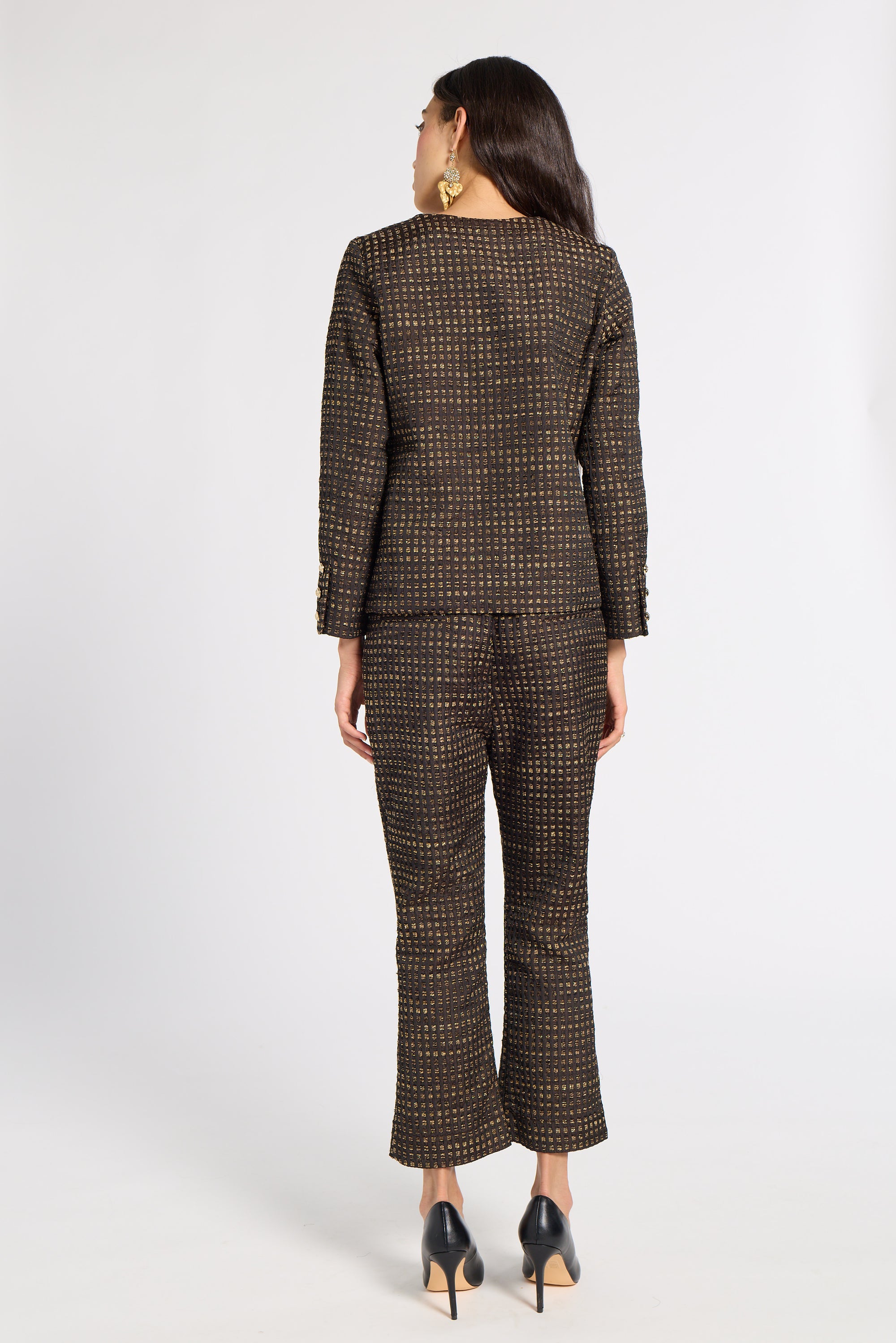 A woman shown from the back wearing a matching dark brown tweed blazer and cropped trousers set with a small gold geometric pattern, paired with black high heels, against a light gray studio background.