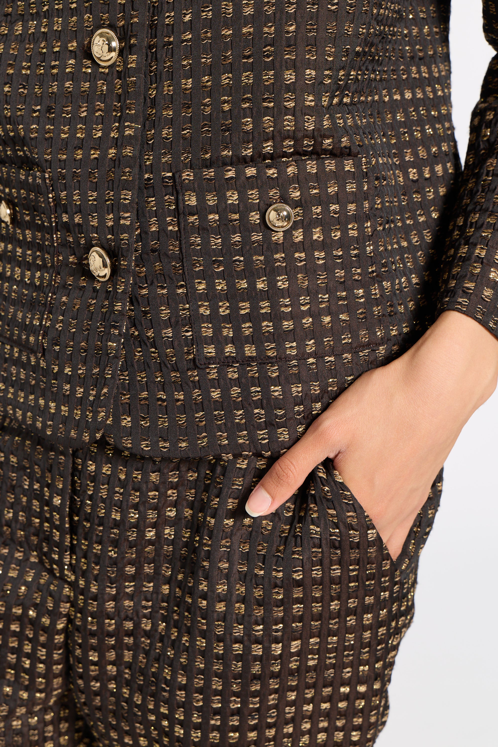 Close-up of a textured black and gold tweed jacket with gold buttons, featuring a hand tucked into a side pocket.
