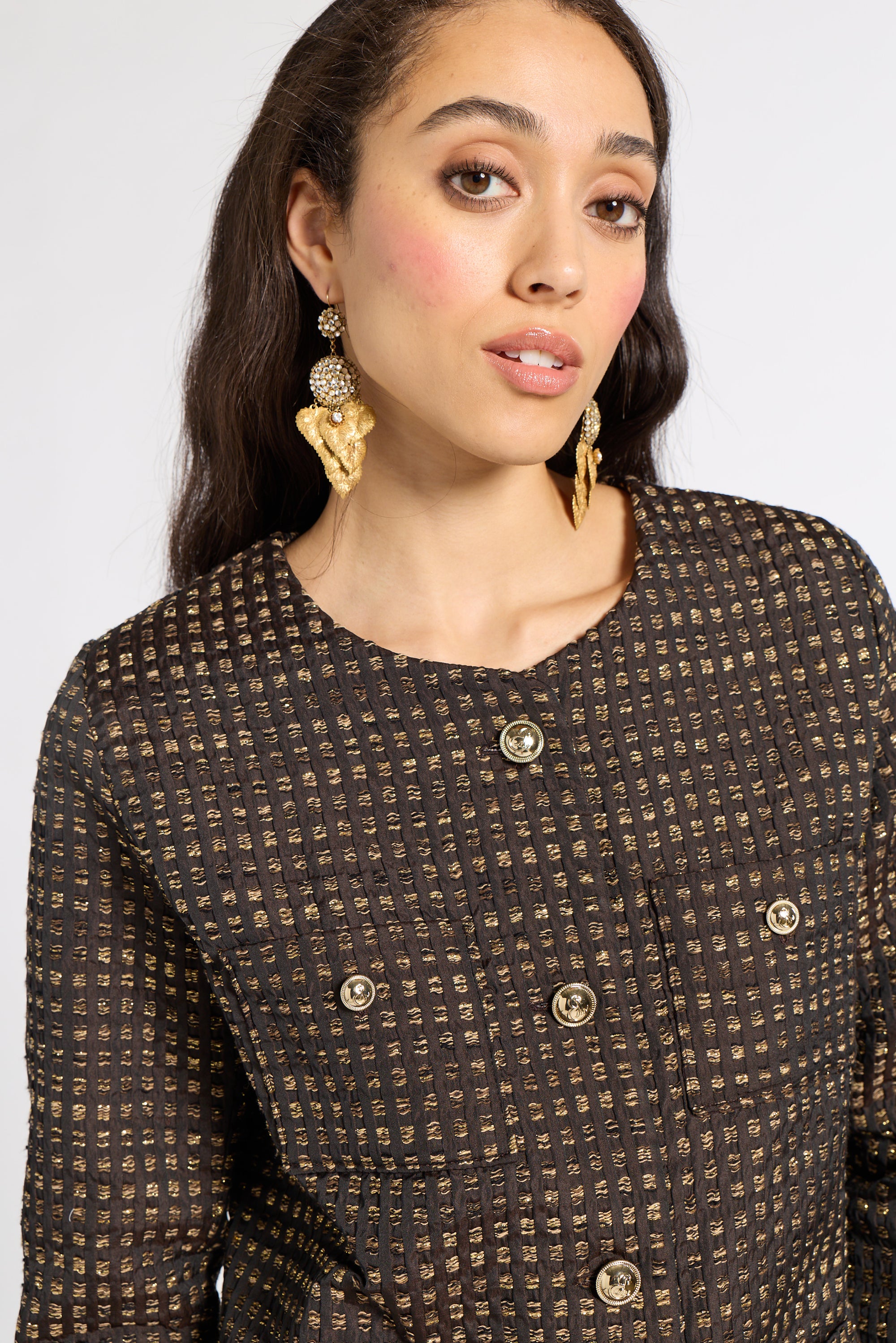A close-up of a woman wearing a dark brown tweed blazer with gold buttons and matching gold statement earrings against a light gray background.