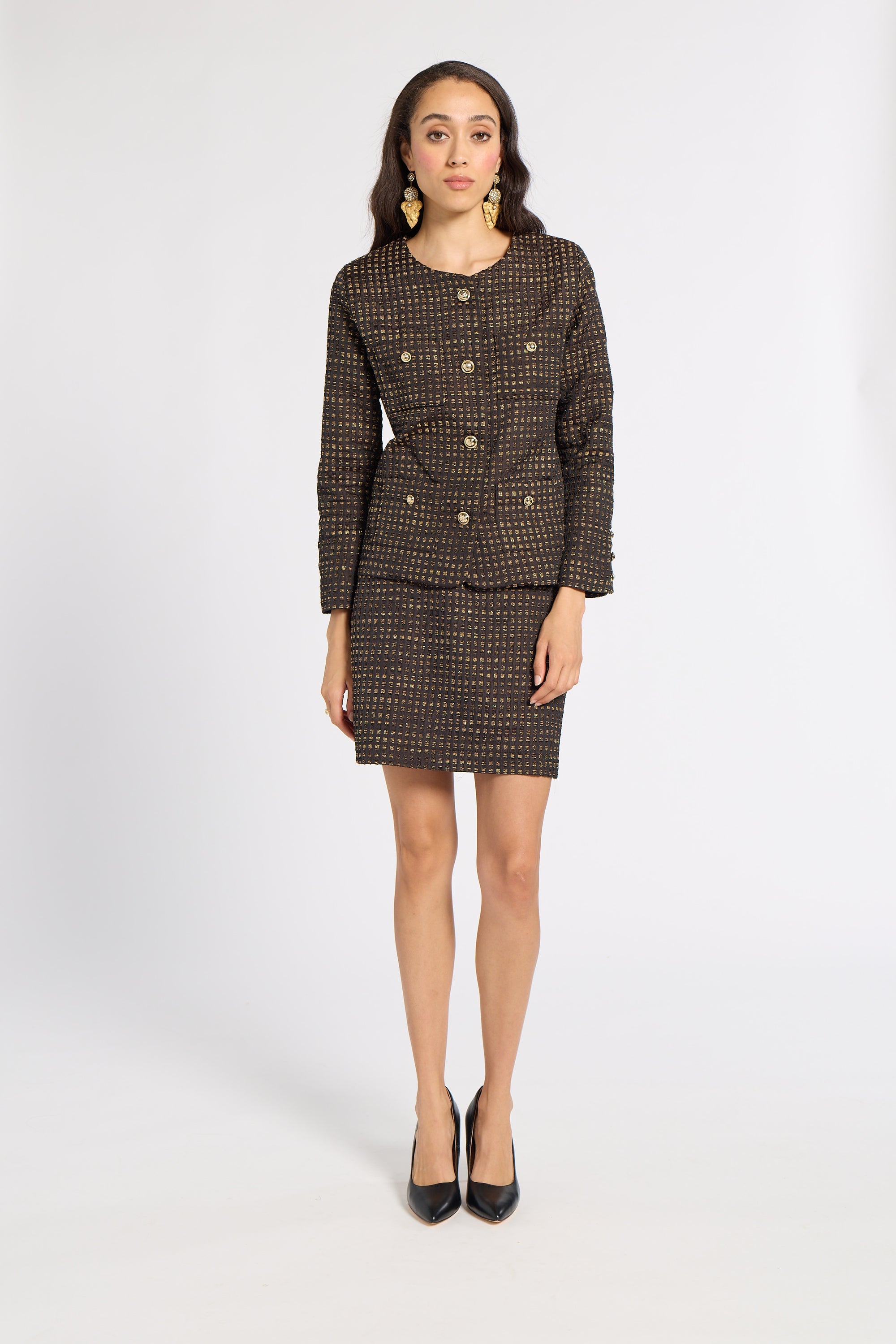 A woman models a matching two-piece tweed suit set consisting of a double-breasted blazer with gold buttons and a matching mini skirt, in a brown and beige micro-pattern. She is wearing black pointed-toe heels and gold statement earrings against a plain white studio background.