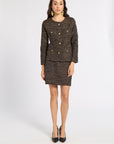 A woman models a matching two-piece tweed suit set consisting of a double-breasted blazer with gold buttons and a matching mini skirt, in a brown and beige micro-pattern. She is wearing black pointed-toe heels and gold statement earrings against a plain white studio background.