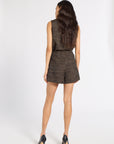 A woman viewed from the back wearing a sleeveless black romper with a textured gold geometric pattern, paired with black high heels, against a plain white studio background.