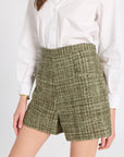 A model wearing high-waisted tweed shorts in an olive green and beige pattern, paired with a white button-down shirt, against a plain light gray background.