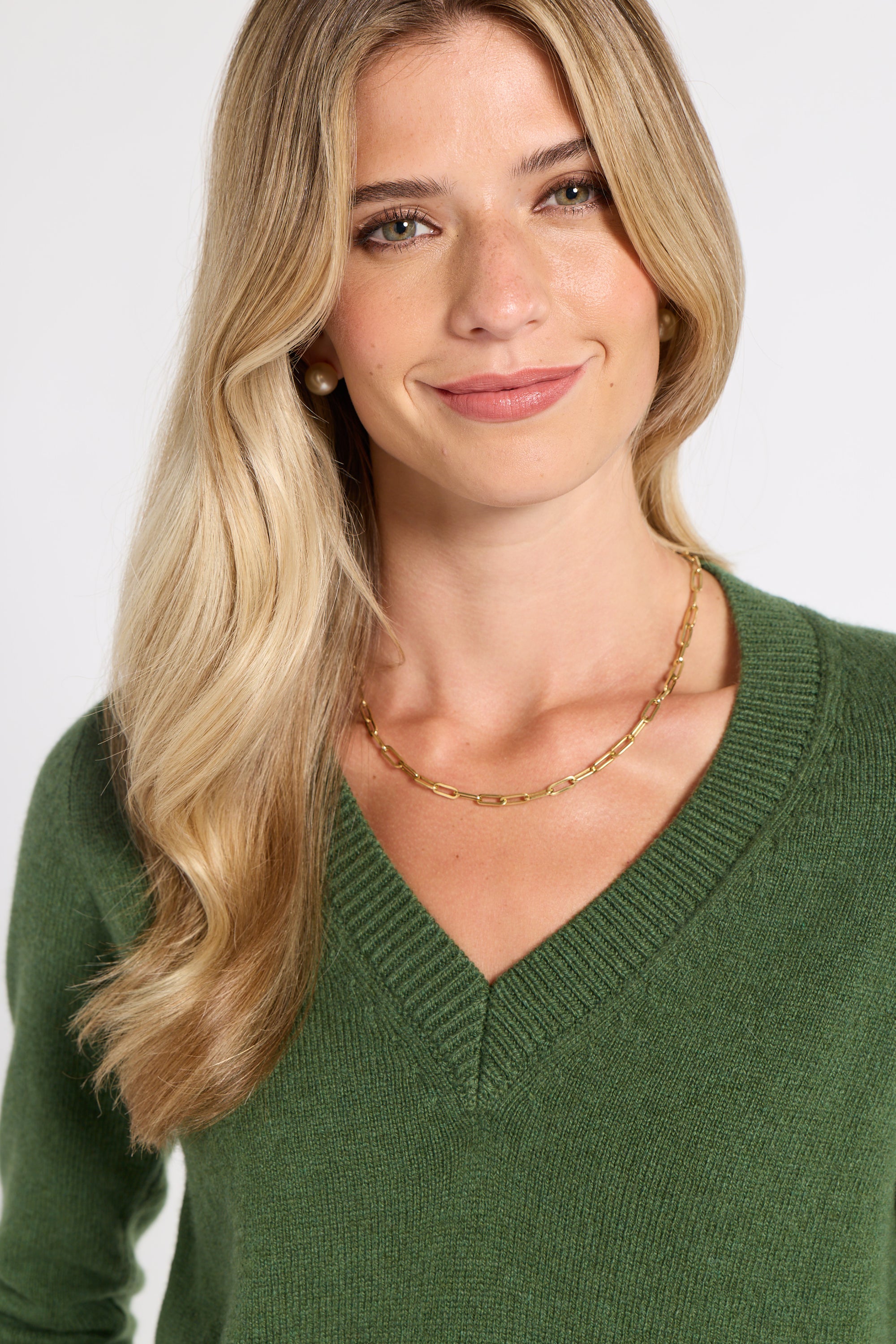 A blonde woman wearing a green V-neck knit sweater and a delicate gold chain necklace, shown from the chest up against a plain light background.