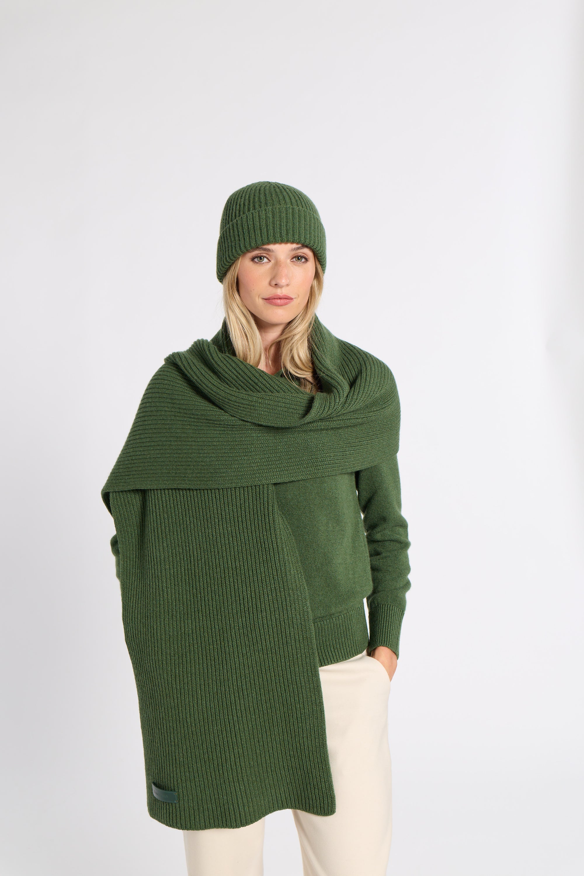 A woman wearing a dark green ribbed knit beanie, matching green wrap shawl, and cream-colored pants, standing against a plain light gray studio background.