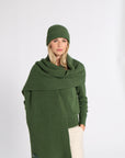 A woman wearing a dark green ribbed knit beanie, matching green wrap shawl, and cream-colored pants, standing against a plain light gray studio background.