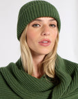 A woman wearing a dark green ribbed knit beanie and a matching green ribbed knit cowl scarf, photographed against a plain light gray studio background.