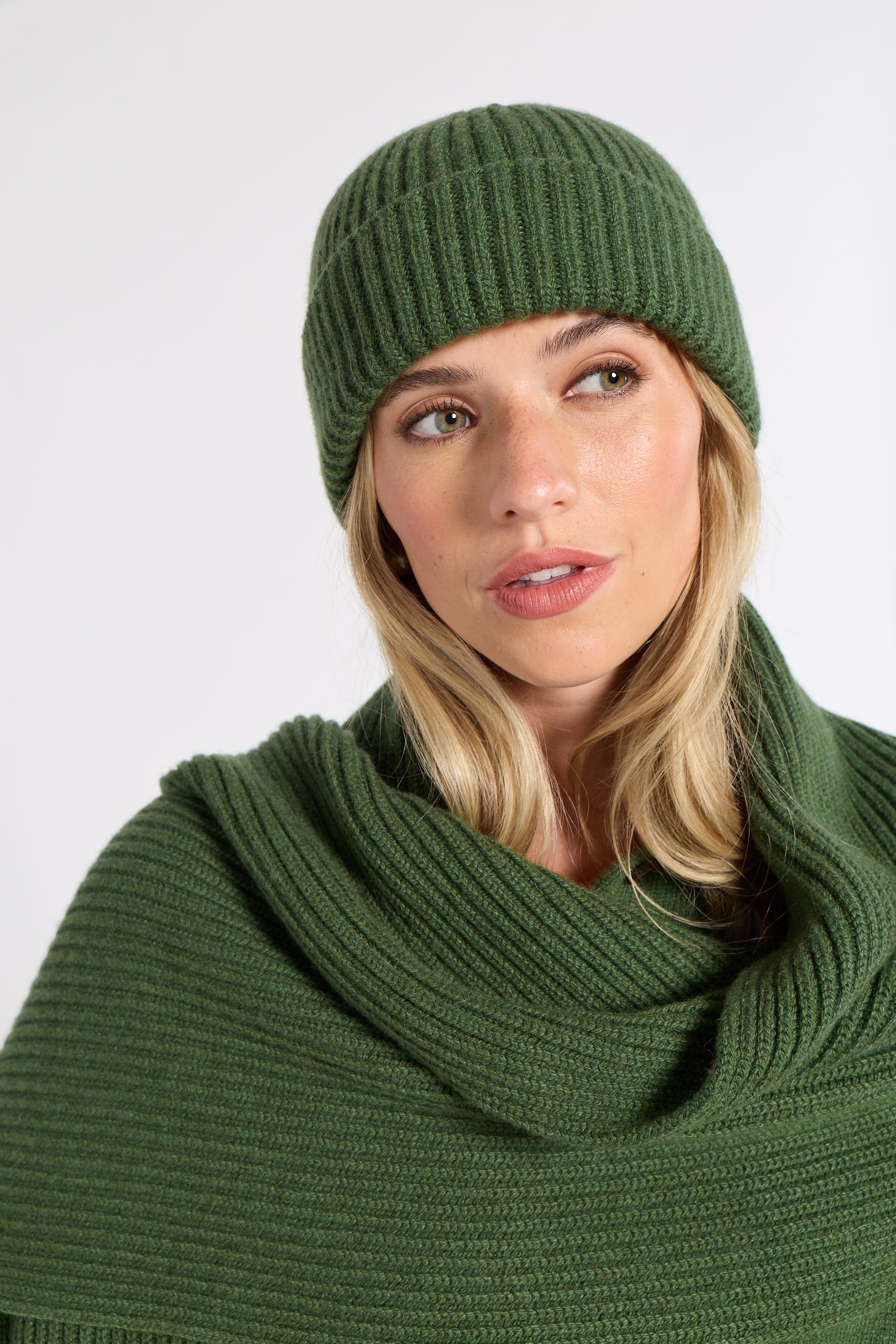 A woman wearing a ribbed knit beanie and matching cowl neck scarf in olive green, photographed against a plain light gray studio background.