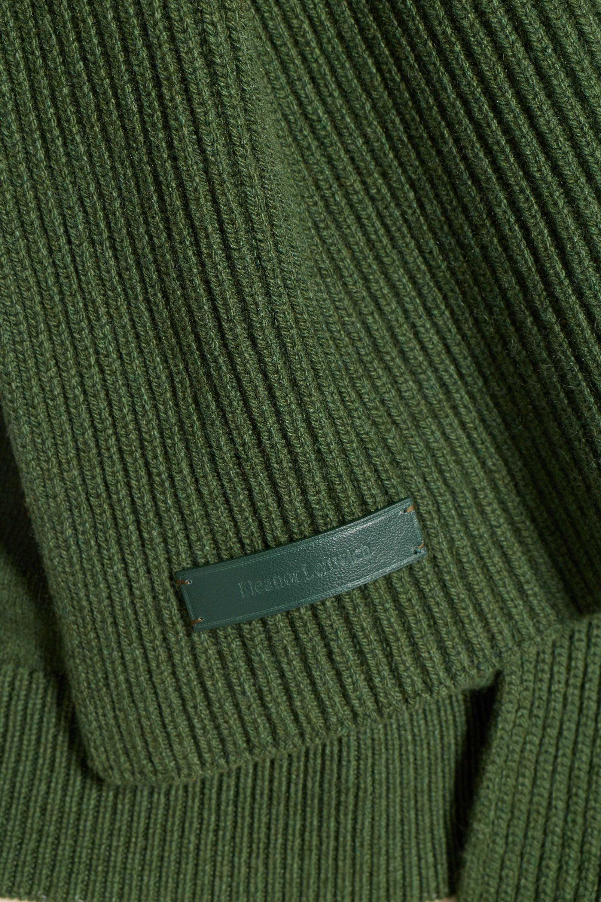 Close-up of a ribbed olive green knit garment with a small dark green leather brand tab reading 'Fleury's Army Co.'