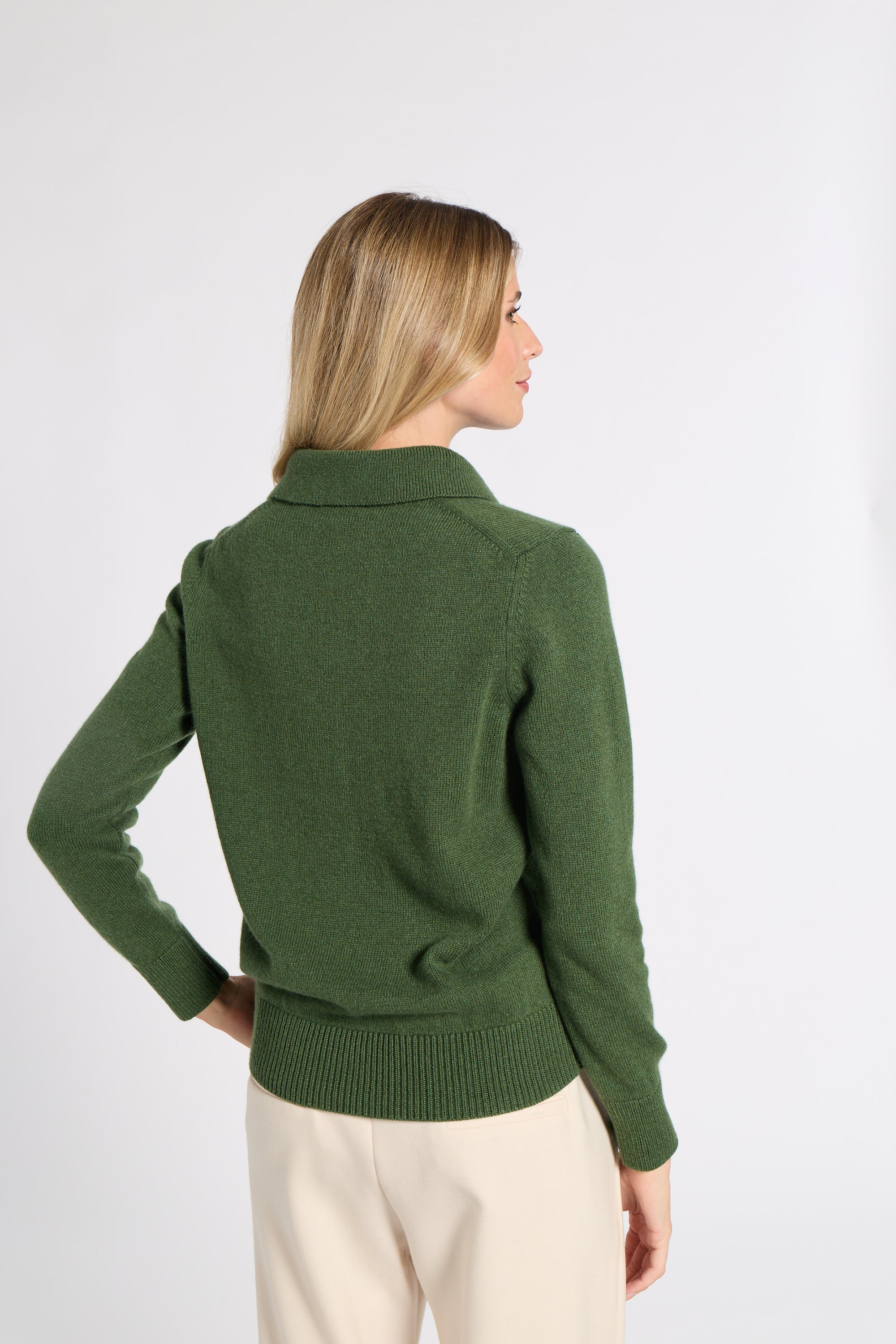 A woman shown from the back wearing a dark green knit sweater with a mock neck and cream-colored trousers, set against a plain light gray studio background.