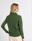 A woman shown from the back wearing a dark green knit sweater with a mock neck and cream-colored trousers, set against a plain light gray studio background.
