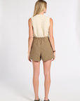 A woman viewed from behind wearing a sleeveless cream cable-knit top and brown plaid shorts, standing against a plain light gray studio background.