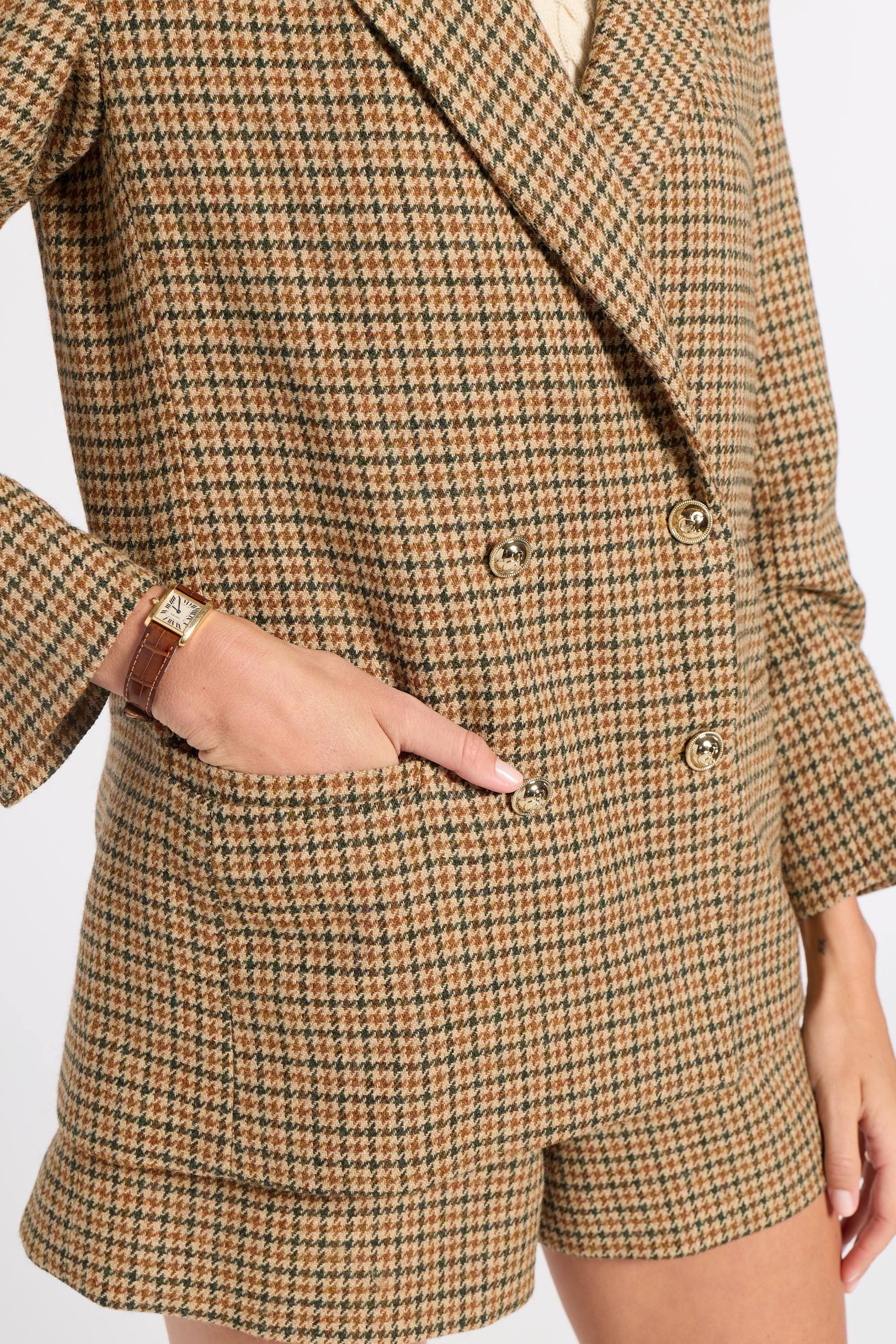 A close-up of a person wearing a brown and beige houndstooth blazer and matching shorts, with one hand in a pocket and a gold watch visible on the wrist.