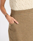 Close-up of a person wearing a brown and beige houndstooth skirt with a side pocket, paired with a cream cable-knit top. The person's hand is in the pocket, and a gold watch is visible on the wrist, against a plain white background.