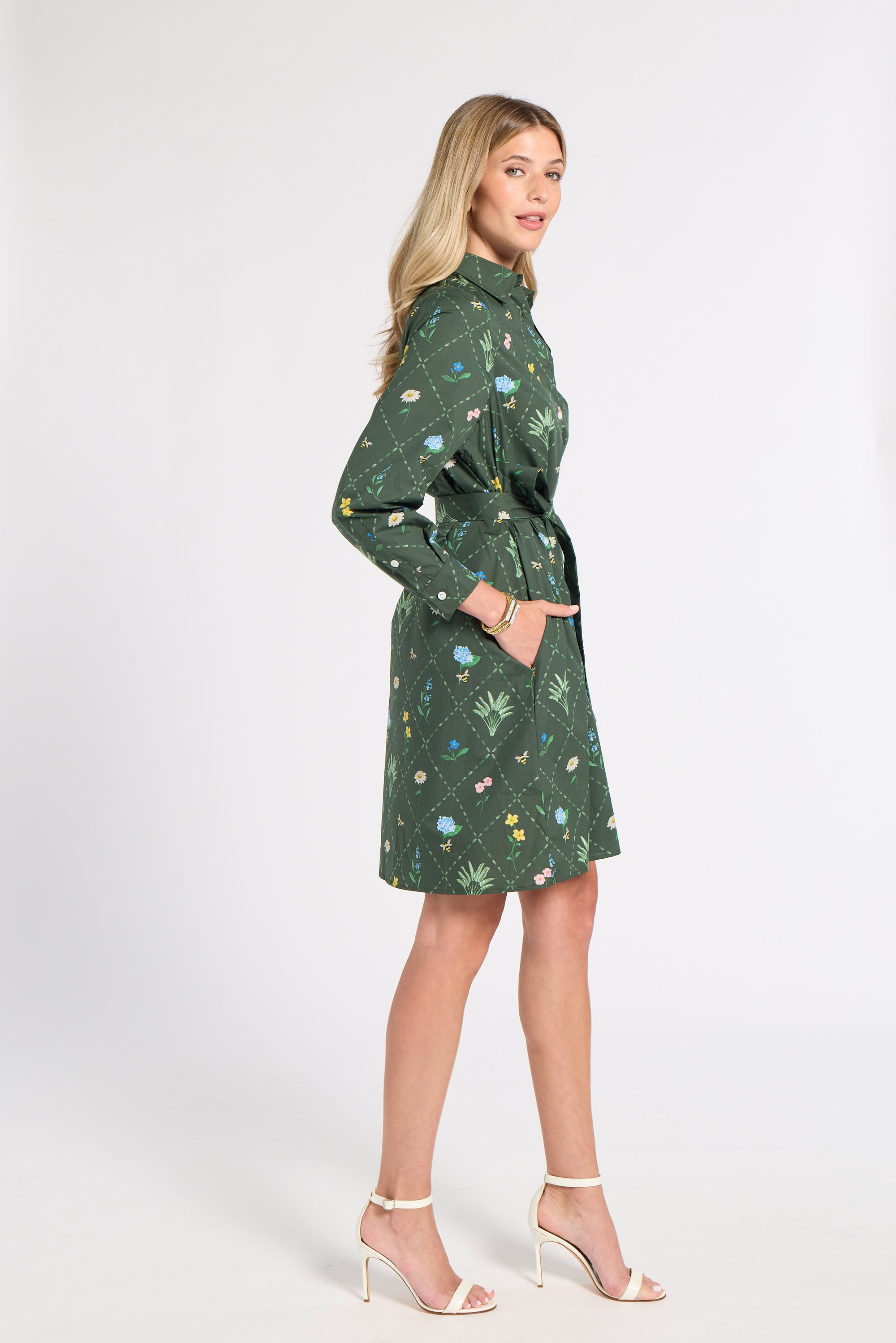 A woman modeling a green long-sleeve mini dress with a botanical print and a matching tie belt, standing against a plain light gray studio background.