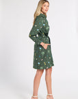 A woman modeling a green long-sleeve mini dress with a botanical print and a matching tie belt, standing against a plain light gray studio background.