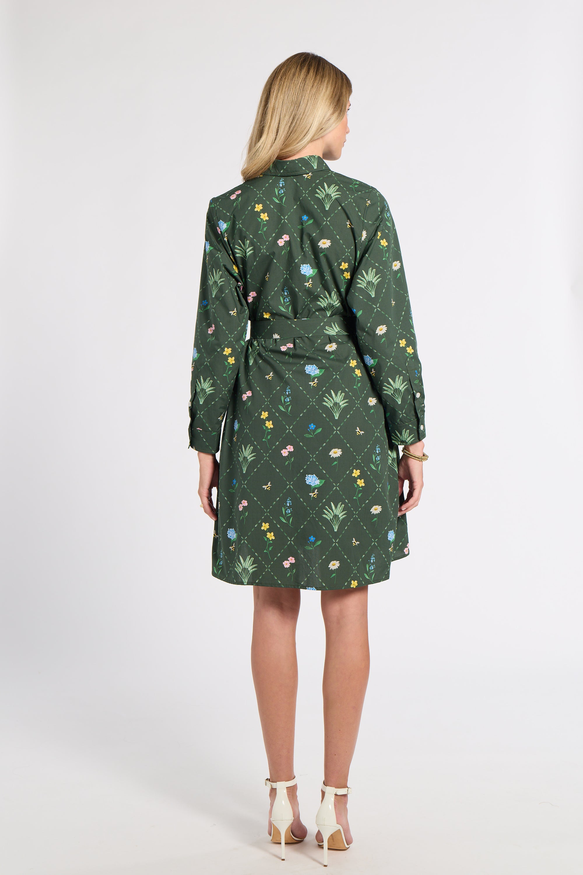 A woman viewed from the back wearing a dark green long-sleeve shirt dress with a colorful floral and botanical print, paired with white high-heeled sandals, against a plain light gray studio background.