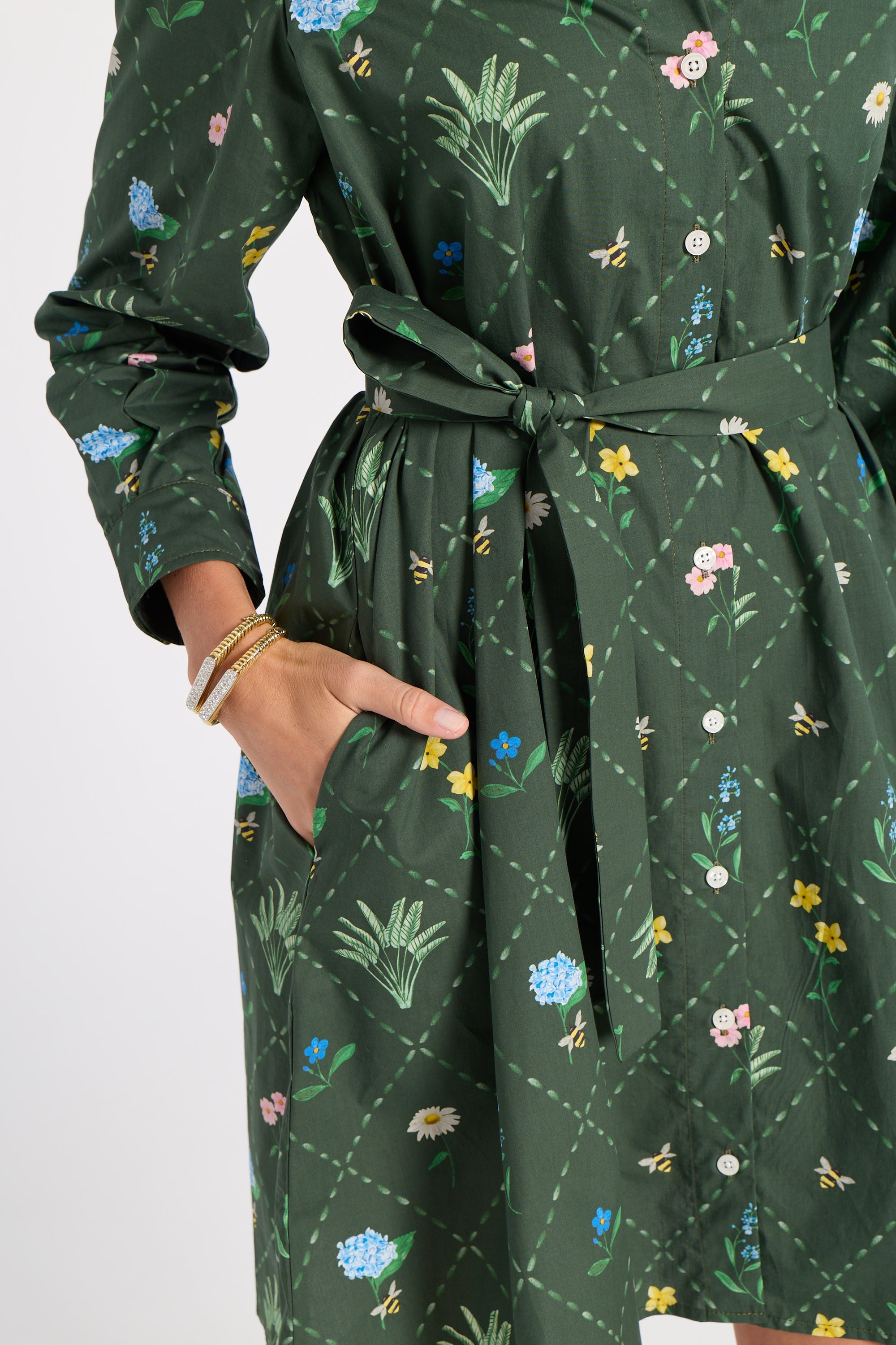 A dark green long-sleeve dress with a tie waist and a colorful floral and bee pattern, shown on a model with a hand in the pocket.