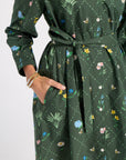 A dark green long-sleeve dress with a tie waist and a colorful floral and bee pattern, shown on a model with a hand in the pocket.