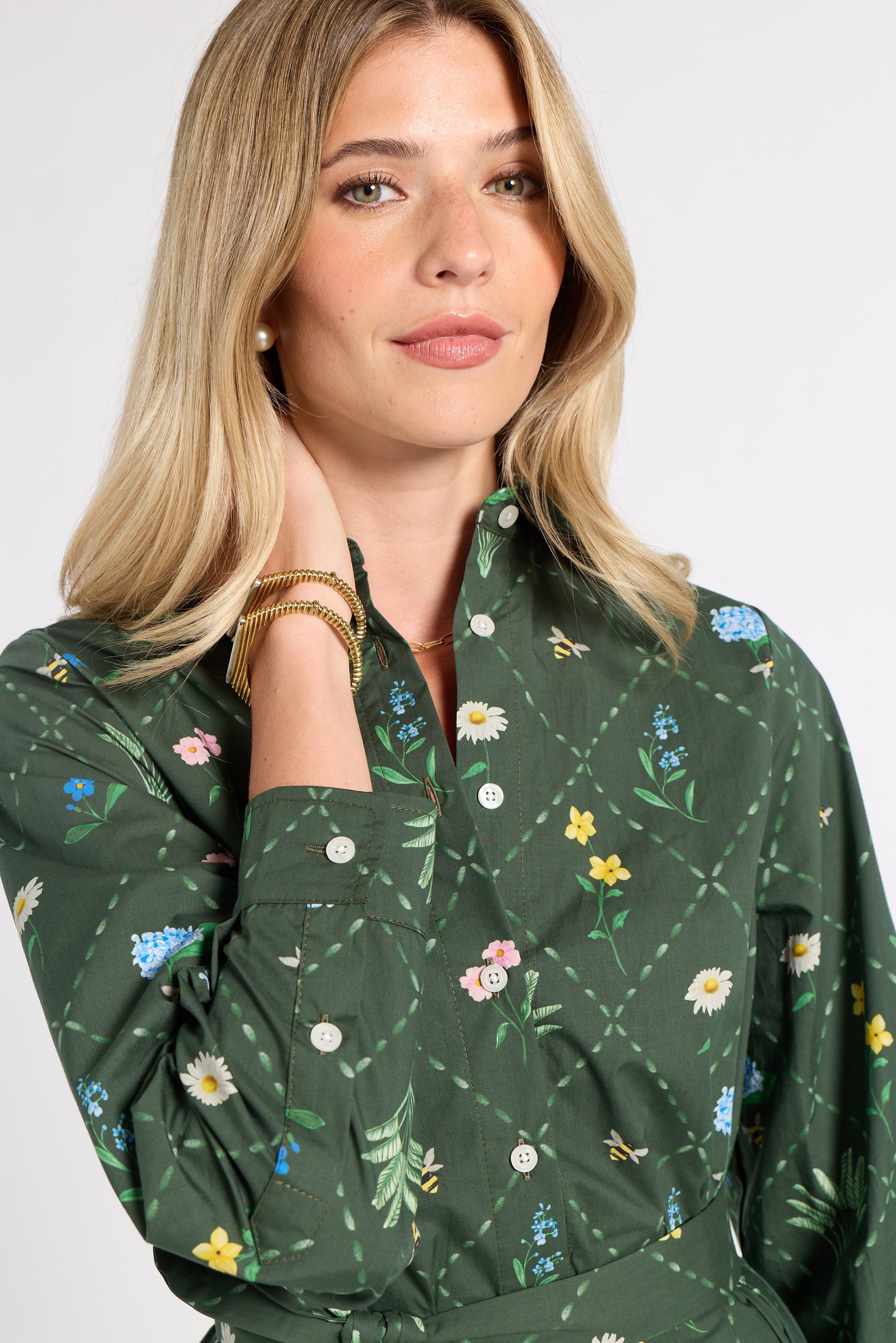 A woman wearing a dark green long-sleeve button-up shirt with a colorful floral and bee pattern, posed against a light gray studio background.