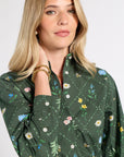 A woman wearing a dark green long-sleeve button-up shirt with a colorful floral and bee pattern, posed against a light gray studio background.