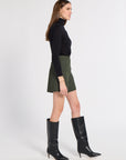 A woman modeling a black long-sleeve turtleneck top, an olive green mini skirt, and black knee-high leather boots, shown in profile against a plain white studio background.