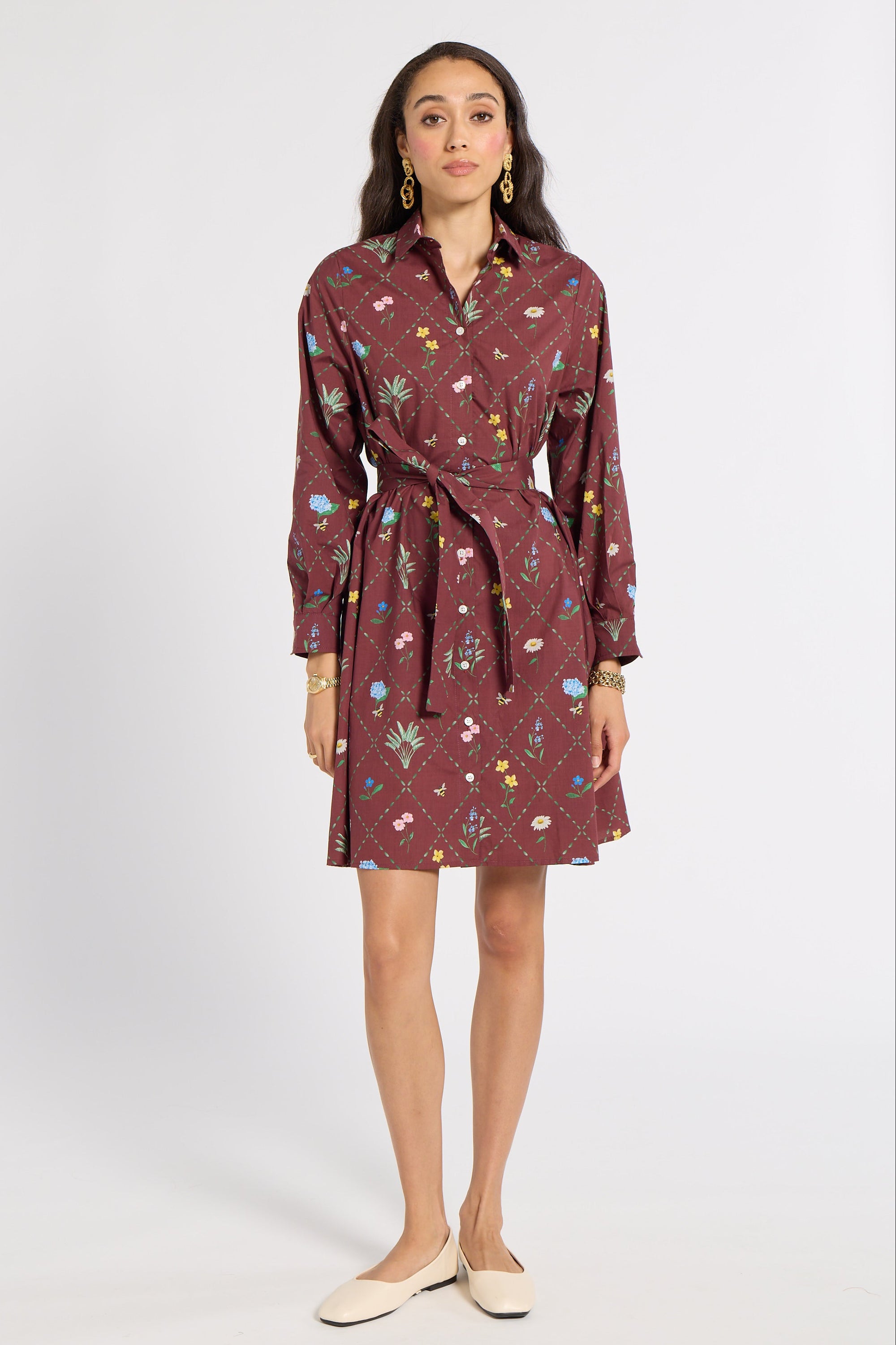 A woman modeling a long-sleeve, collared wrap dress with a tie waist. The dress has a dark reddish-brown base color with a colorful floral and botanical print.