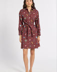 A woman modeling a long-sleeve, collared wrap dress with a tie waist. The dress has a dark reddish-brown base color with a colorful floral and botanical print.
