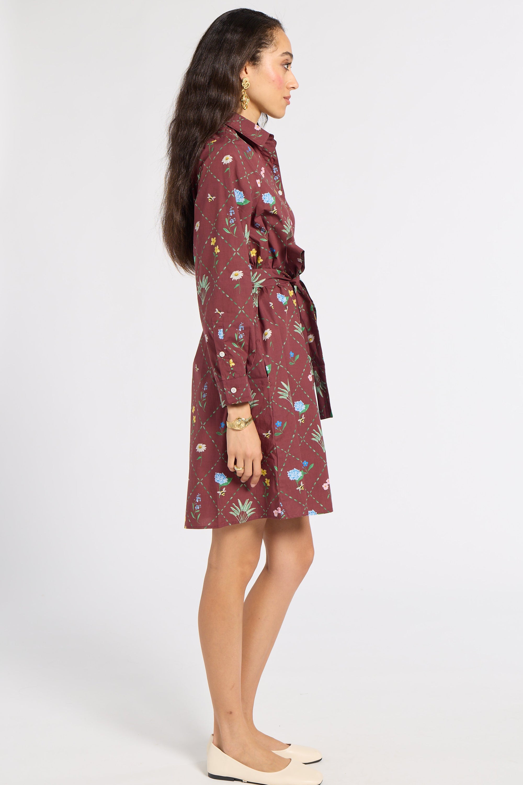 A woman modeling a long-sleeve, belted shirt dress in a maroon color with a colorful floral and botanical print, shown in profile against a plain white studio background.