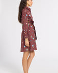 A woman modeling a long-sleeve, belted shirt dress in a maroon color with a colorful floral and botanical print, shown in profile against a plain white studio background.