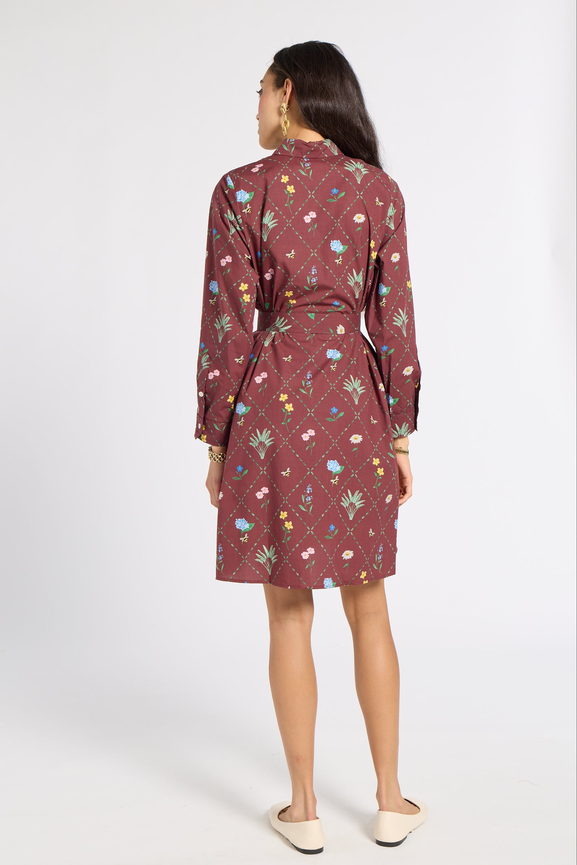 A woman is shown from the back wearing a long-sleeved, knee-length dress with a multicolor floral and botanical pattern on a dusty rose base, paired with beige flat shoes against a white studio background.