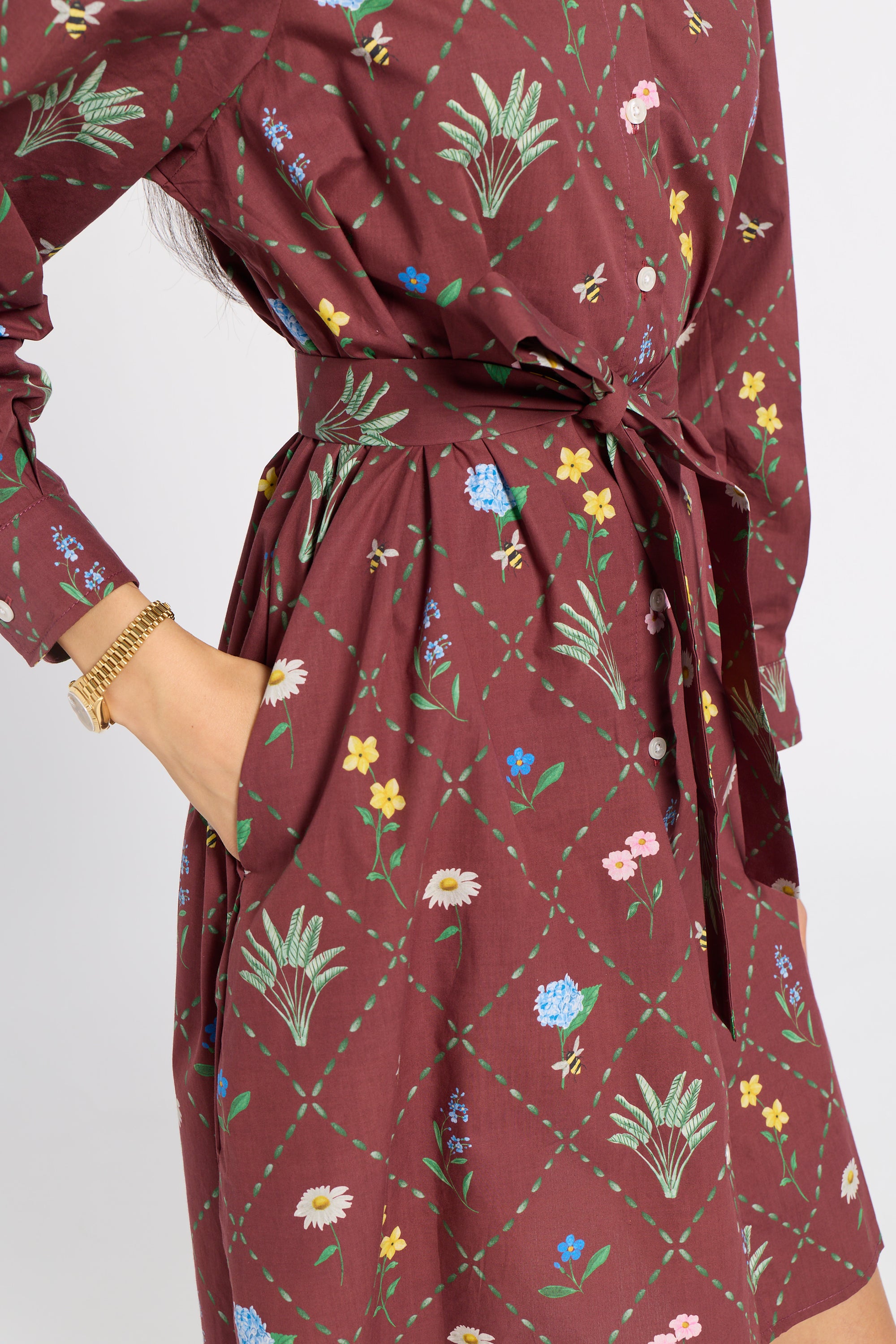 A close-up of a woman wearing a long-sleeve wrap dress in a deep burgundy color with a green diamond lattice pattern and colorful floral prints. The dress features a tie waist and side pockets, and the model is wearing a gold chain bracelet.