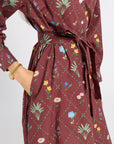 A close-up of a woman wearing a long-sleeve wrap dress in a deep burgundy color with a green diamond lattice pattern and colorful floral prints. The dress features a tie waist and side pockets, and the model is wearing a gold chain bracelet.