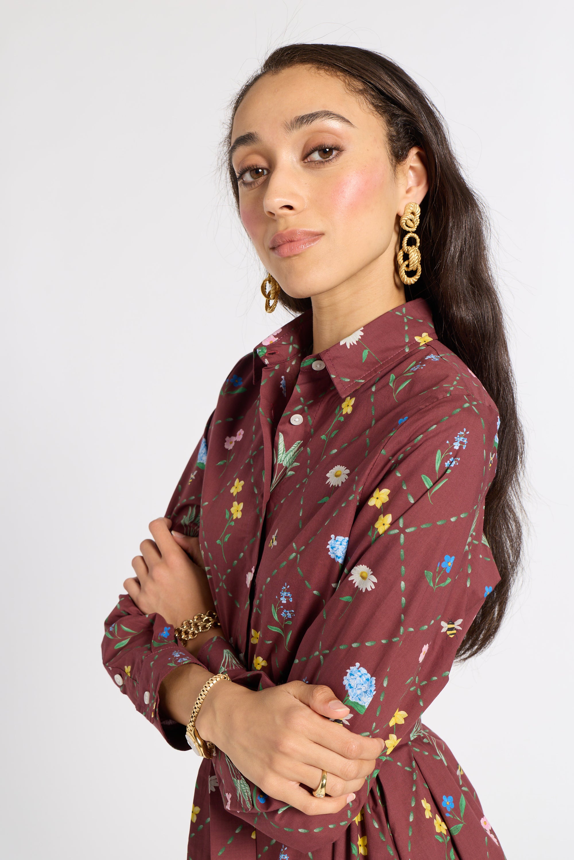 A woman modeling a long-sleeve collared shirt dress in maroon with a colorful floral and vine print, accessorized with gold jewelry against a plain light gray background.