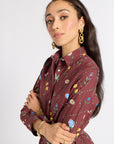A woman modeling a long-sleeve collared shirt dress in maroon with a colorful floral and vine print, accessorized with gold jewelry against a plain light gray background.