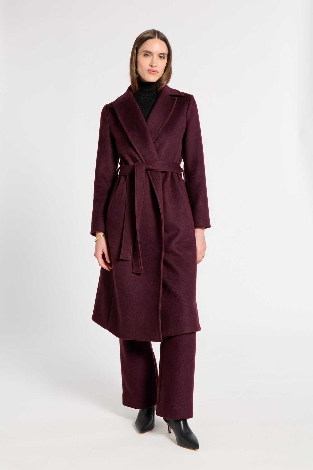 A woman modeling a long burgundy wrap coat with a matching waist tie, paired with matching wide-leg trousers, a black turtleneck, and black pointed-toe heels, against a plain light gray studio background.