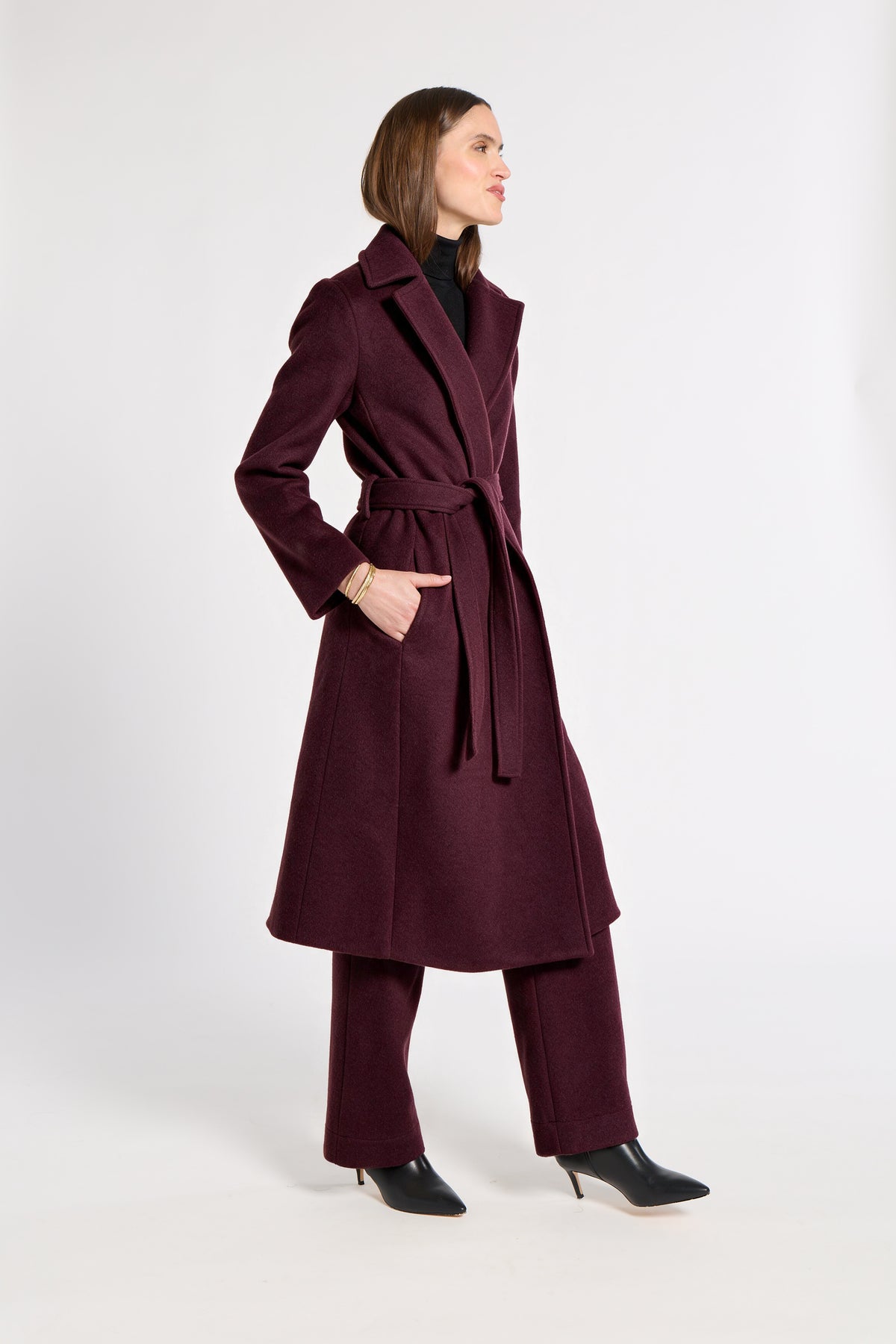 A woman modeling a long, belted  cashmere coat in deep burgundy, paired with matching trousers and black pointed-toe heels, photographed against a plain light gray studio background.