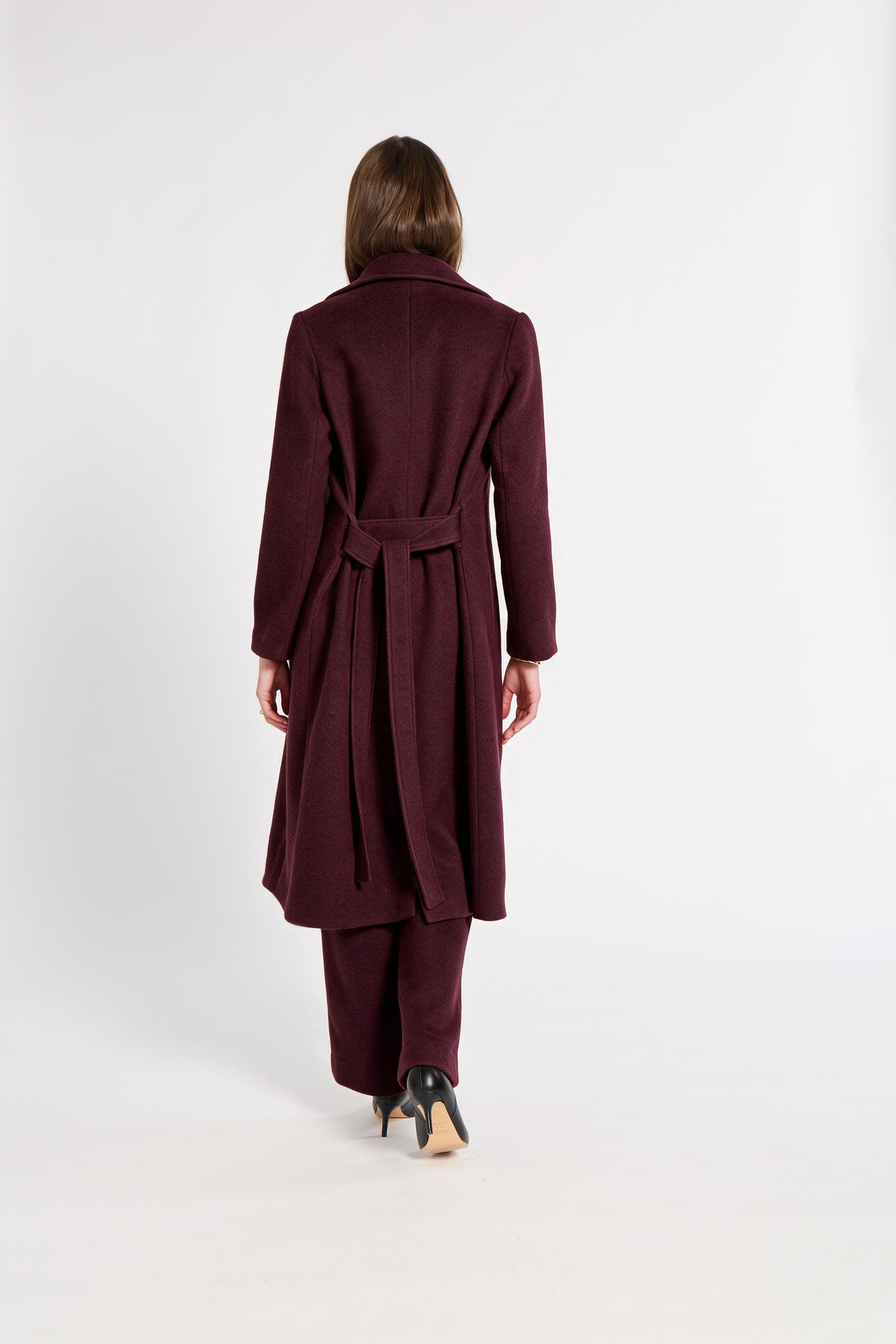 A woman viewed from behind wearing a long burgundy cashmere belted coat and matching trousers against a plain white studio background.