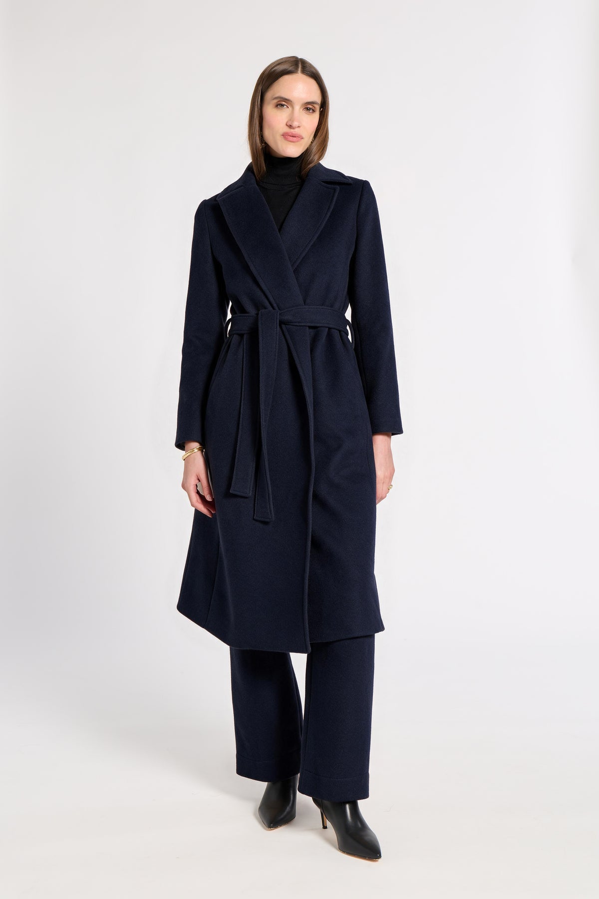 A woman modeling a long navy wrap-style coat with a matching waist tie, worn over a black turtleneck and black wide-leg trousers, against a plain light gray studio background.