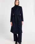 A woman modeling a long navy wrap-style coat with a matching waist tie, worn over a black turtleneck and black wide-leg trousers, against a plain light gray studio background.